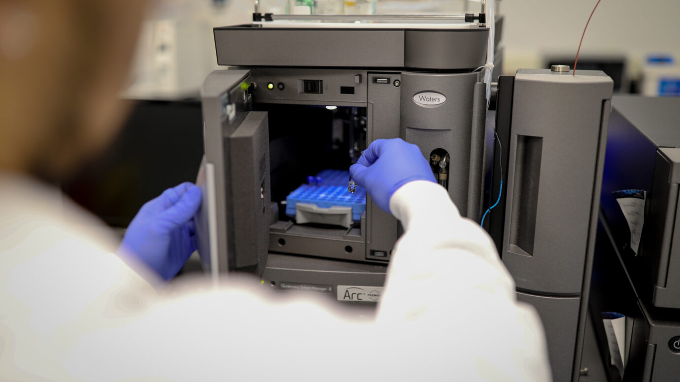 Gloved hands loading a sample tray into a Waters laboratory analyzer