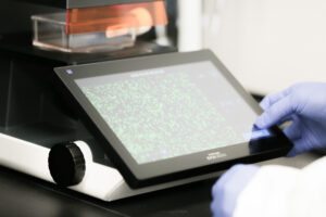 Gloved hand holding a sample cassette next to a lab instrument display showing a DNA sequencing chromatogram
