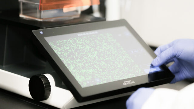 Gloved hand holding a sample cassette next to a lab instrument display showing a DNA sequencing chromatogram