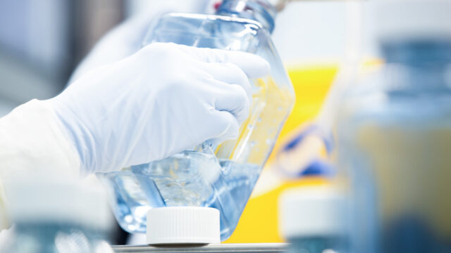 Gloved hand holding a bottle of orange liquid in a manufacturing cleanroom
