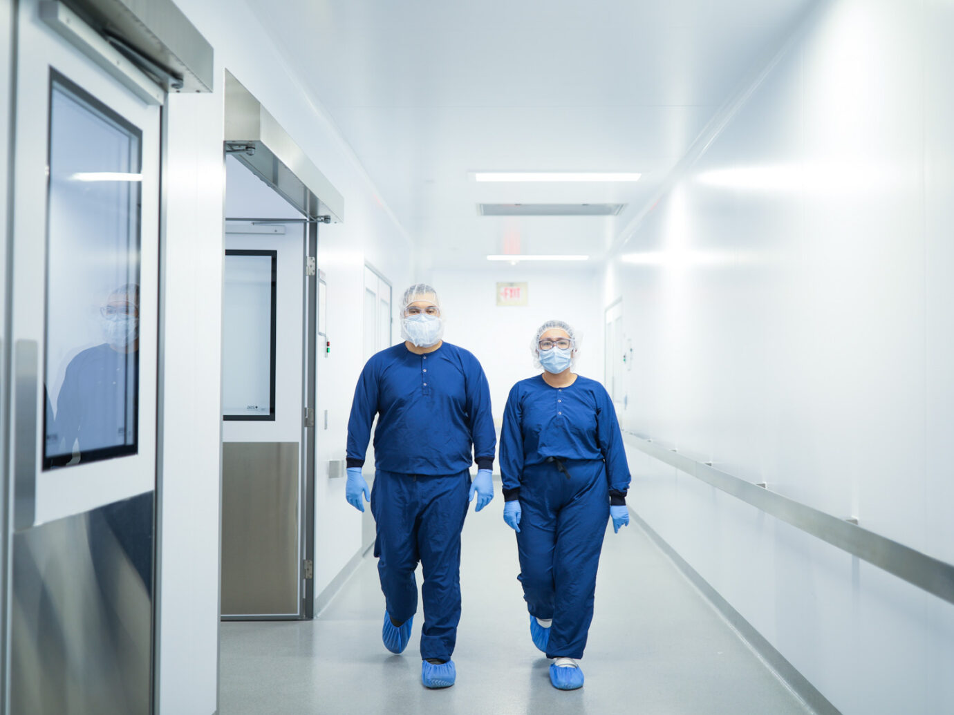 Two employees in blue scrubs, hairnets, and masks standing in a facility hallway
