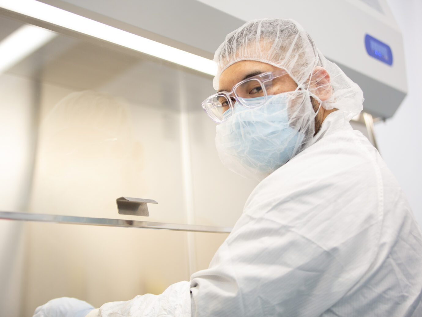 Worker in a mask, goggles, and hairnet looking into a cleanroom biosafety cabinet