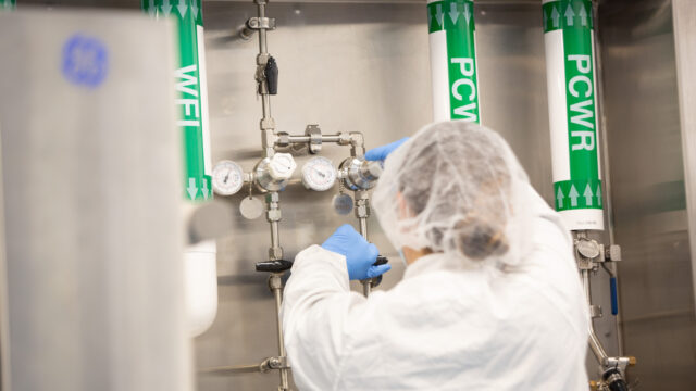 Technician in a hairnet adjusting valves on labeled gas supply lines in a manufacturing suite