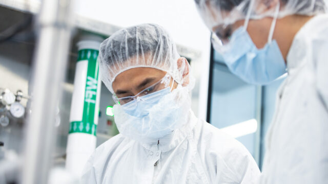 Two cleanroom workers in masks and hairnets standing beside vials on a rack