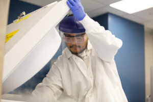 Lab worker in a cleanroom bunny suit and goggles reaching into an overhead storage unit