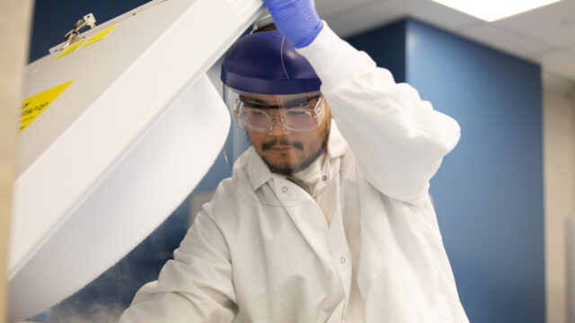 Lab worker in a cleanroom bunny suit and goggles reaching into an overhead storage unit