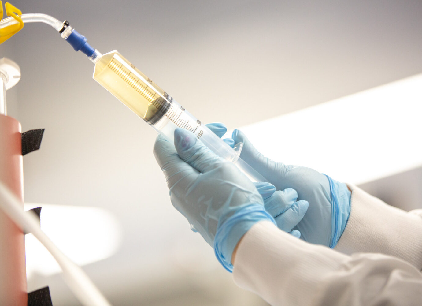 Gloved hand holding a syringe filled with yellow liquid connected to lab tubing