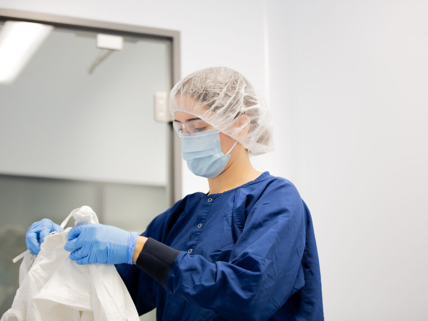 Close-up of a worker wearing a hairnet, mask, and blue scrubs putting on gloves