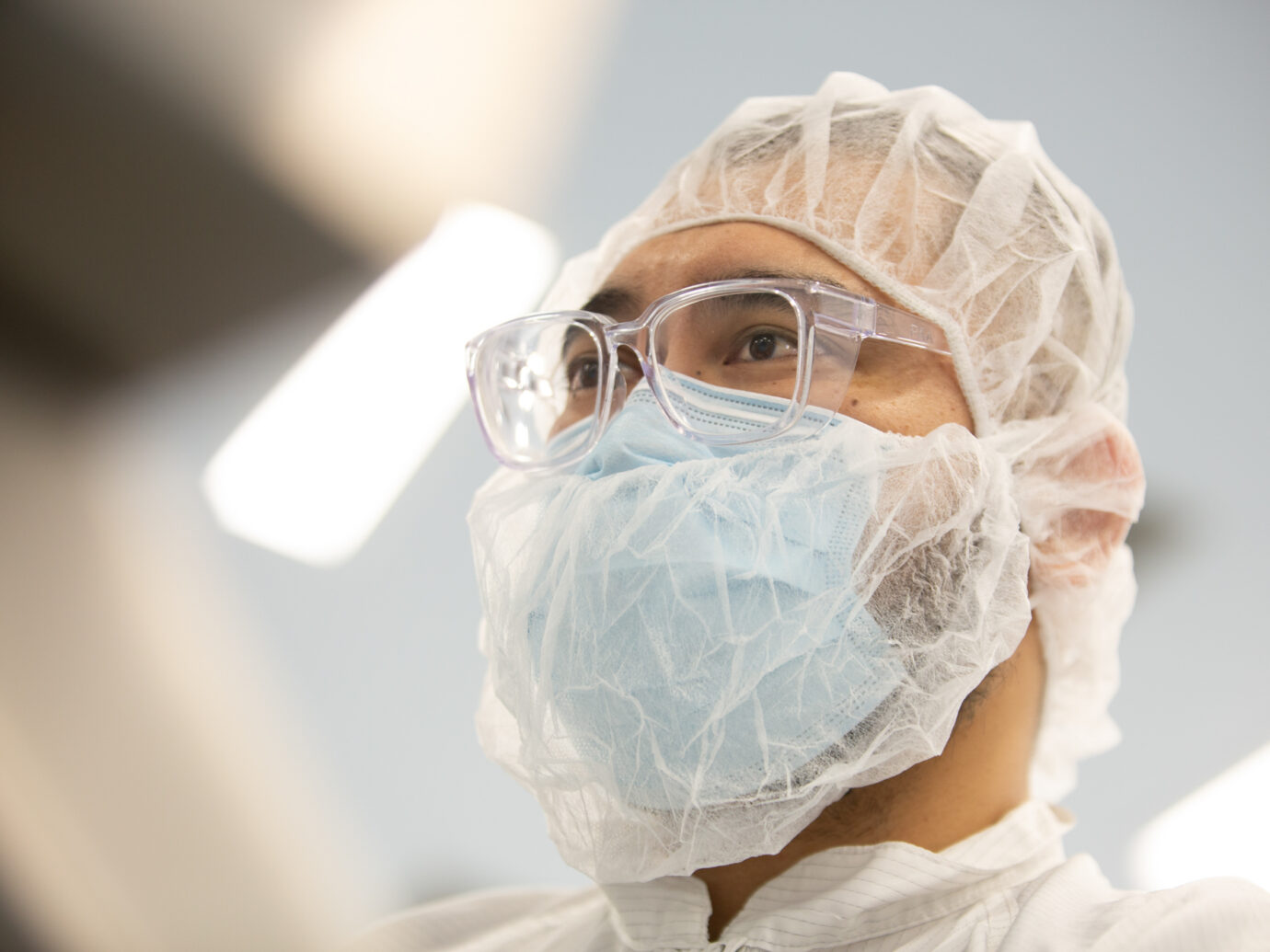 Close-up of a technician wearing a hairnet, safety glasses, and mask in a cleanroom