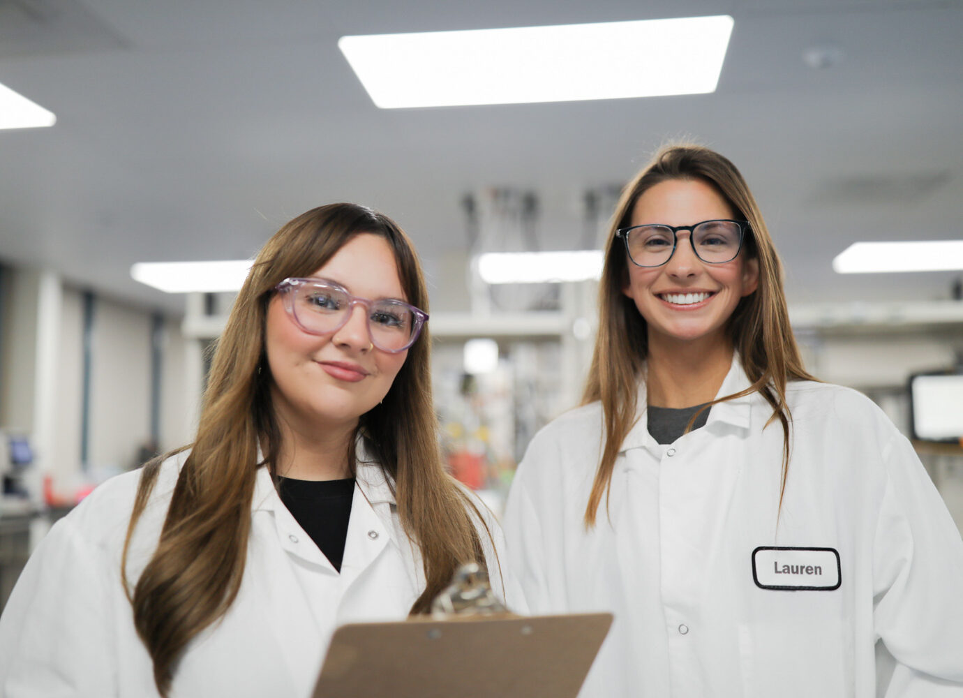 Two smiling female scientists wearing lab coats in a bright office setting